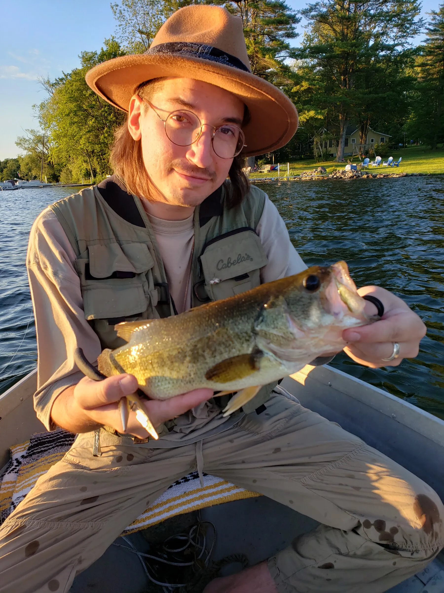 Colton Spahmer on a boat at golden hour, in a wide-brimmed hat and fishing vest, holding a largemouth bass.